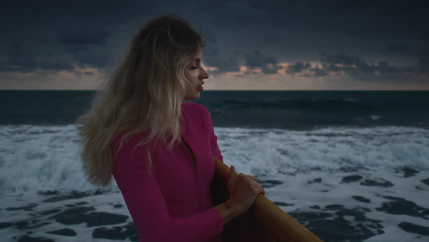 Portrait: beautiful girl with a surfboard. Face of a sexy woman on the shore or beach. Blonde female enjoys the tropical coast of Bali island. Surf board and oink swim suit. Blue hour and sunset