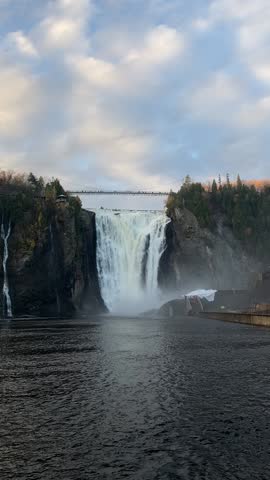 Montmorency Falls near Quebec City, Canada, cascading dramatically from a height of 83 meters with a suspension bridge above, surrounded by autumnal foliage and misty skies