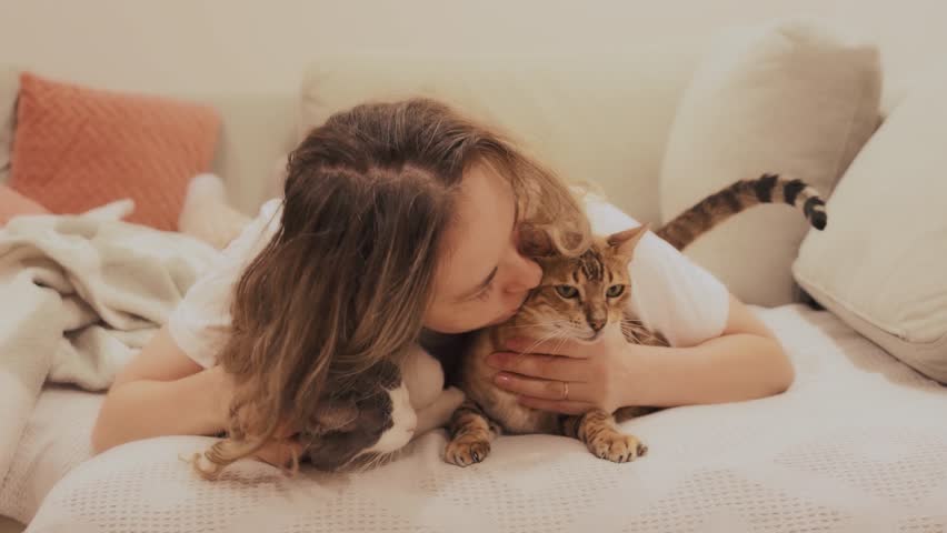 happy woman lying on sofa hugging two cute cats, joyfully kissing bengal cat and non-pedigree fluffy cat. Joyful family person friends with animals pets concept love care. girl casual clothes pajamas.
