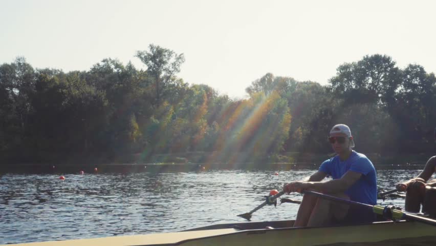 Rowing team training. Side view of 2 young caucasian male rowers, during a rowing practice, athlete sitting in a boat in the river Dnipro, rows through a calm water sunny day, autumn. 4k footage