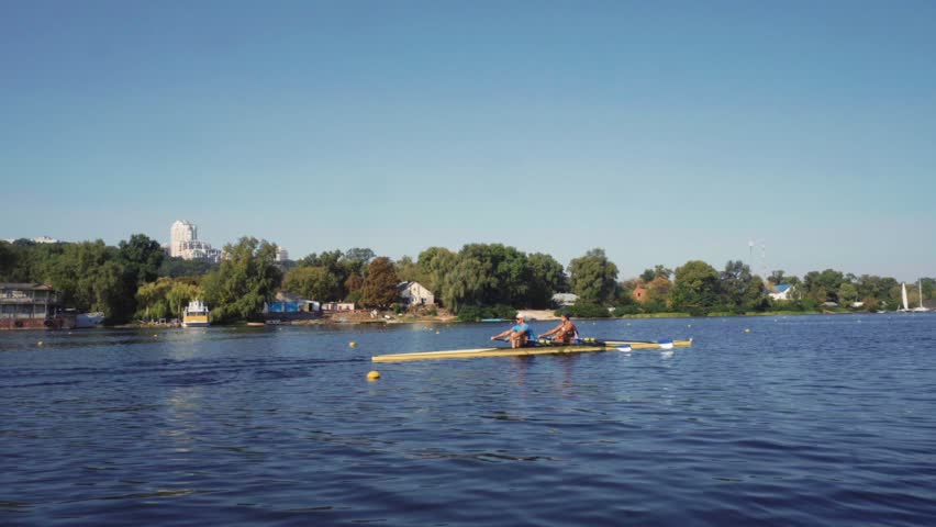 Rowing team training. Side view of 2 young caucasian male rowers, during a rowing practice, athlete sitting in a boat in the river Dnipro, rows through a calm water sunny day, autumn. 4k footage