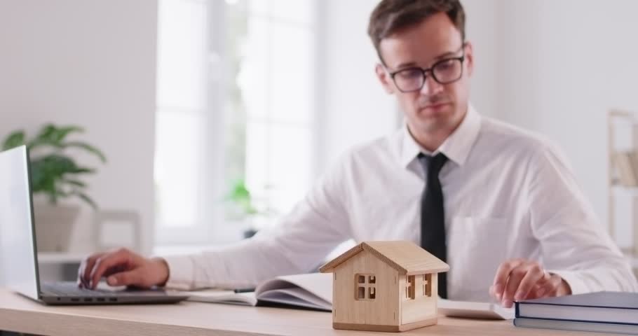 Dedicated real estate agent works at his desk in modern office, immersed in paperwork. Serious young man taking notes while checking his laptop and calculating on calculator. Wooden house is on table.