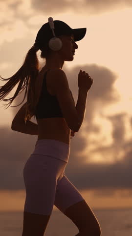 A young sporty woman runner is listening to music in earphones and training. Slim fitness girl is jogging outdoor. Concept of workouts running and healthy sport lifestyle. Vertical video footage