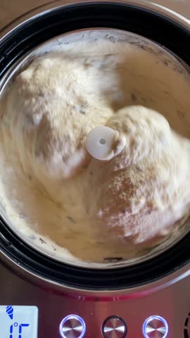 A close-up of ice cream churning in a machine. The mixture is thick and creamy, with visible swirls and a hint of vanilla.