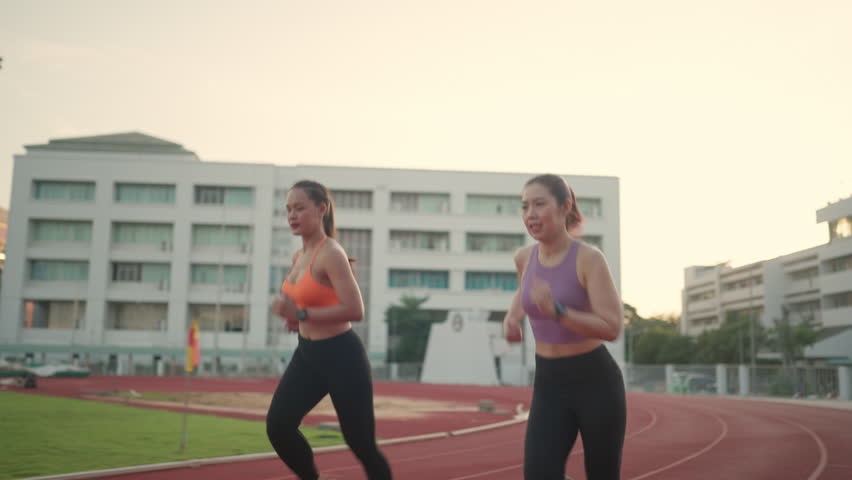 Two women running on a track. One is wearing a pink tank top. The other is wearing an orange top