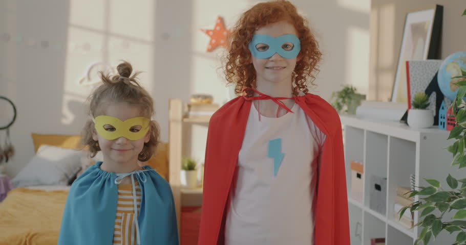 Slow motion portrait of joyful kids siblings wearing costumes masks and capes standing in bedroom raising arms in superhero pose. Childhood and fun concept.