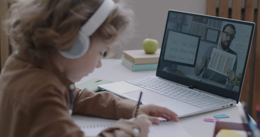 Small child learning English language during online lesson with male teacher looking at laptop screen listening to tutor from home. Education and modern technology concept.