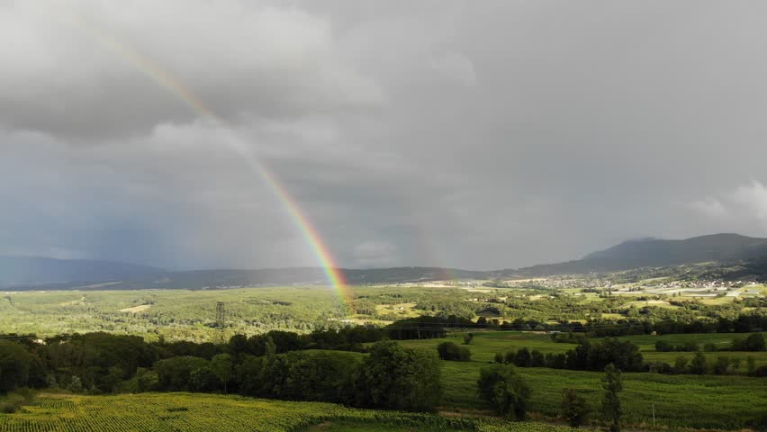 Aerial footage of a rural area in France featuring rolling hills, cultivated fields, a small village, and a vivid rainbow stretching across the sky.
