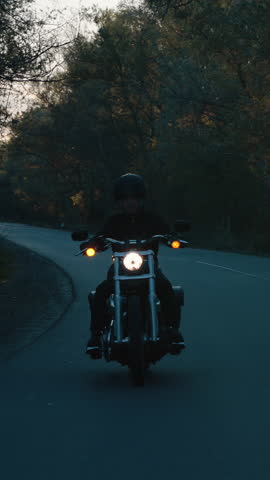 Vertical Screen: Biker in black clothes and helmet riding motorbike in the dark on country road in slow motion, setting sun viewed through trees. Mystical gloomy nature. Concept of adventure