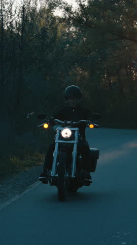 Vertical Screen: Biker in black clothes and helmet riding motorbike in the dark on country road in slow motion, setting sun viewed through trees. Mystical gloomy nature. Concept of adventure