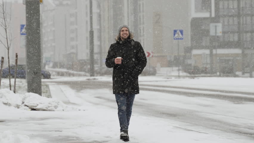 Smiling Caucasian man walking along road in snowfall with cup of hot drink. Portrait of happy and positive person in modern neighborhood in winter. Enjoying snow with passing cars in background.