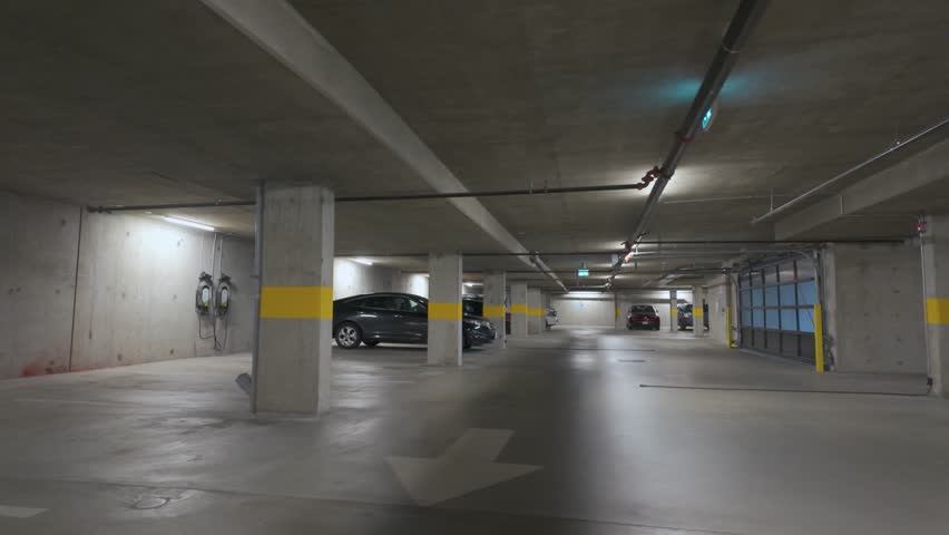 A modern, well-lit underground parking garage. The environment is clean and spacious, with minimal vehicles parked. The lot features prominent parking space markings, directional arrows on the ground
