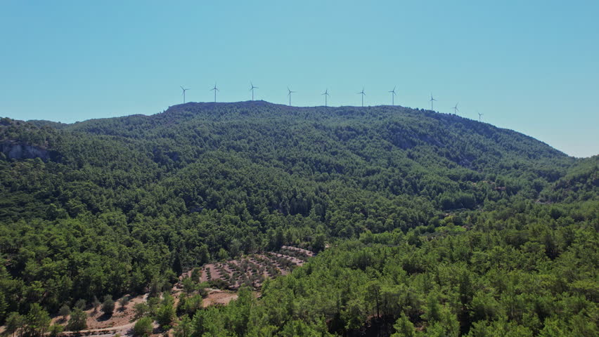 A row of wind turbines stands atop a lush, green mountain under a bright blue sky, surrounded by dense trees and distant rocky formations.