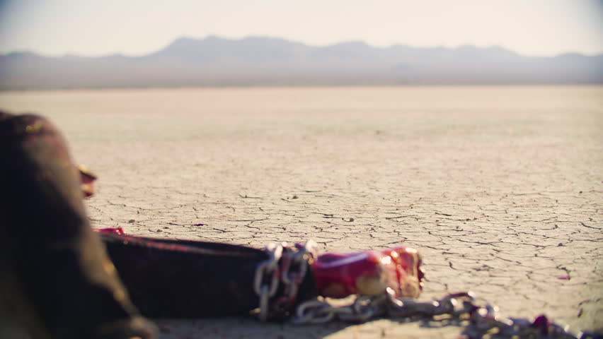 Wounded bleeding man, shackled by the ankle, lies on dry scorched earth, only his legs and feet visible. Enigmatic woman in heavy black boots and flowing shirt steps in and passes by the wounded man