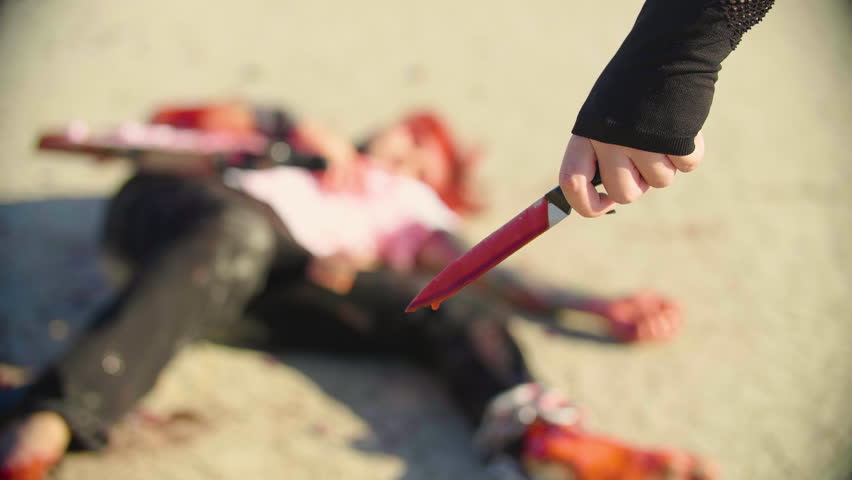 Female hand holds blood-covered knife with blurred figure of a wounded man visible in the background. A mysterious woman with bloody knife stands next to her victim, only her hand and weapon visible.