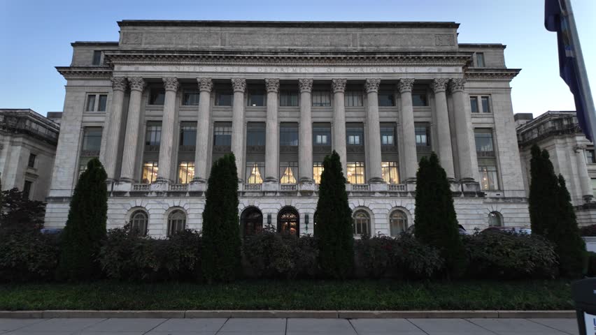A white marble building with columns, windows, and a sign reading United States Department of Agriculture. There are bushes and trees in front of the building. The sky is blue and there is a flagpole 