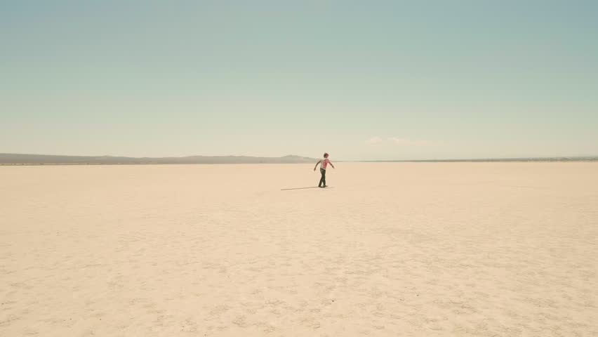 Wide shot of a solitary figure slowly moving through the sands. Lonely man with red dyed hair, punk hairstyle and white t-shirt with blood stains walks slowly through the vast desert.