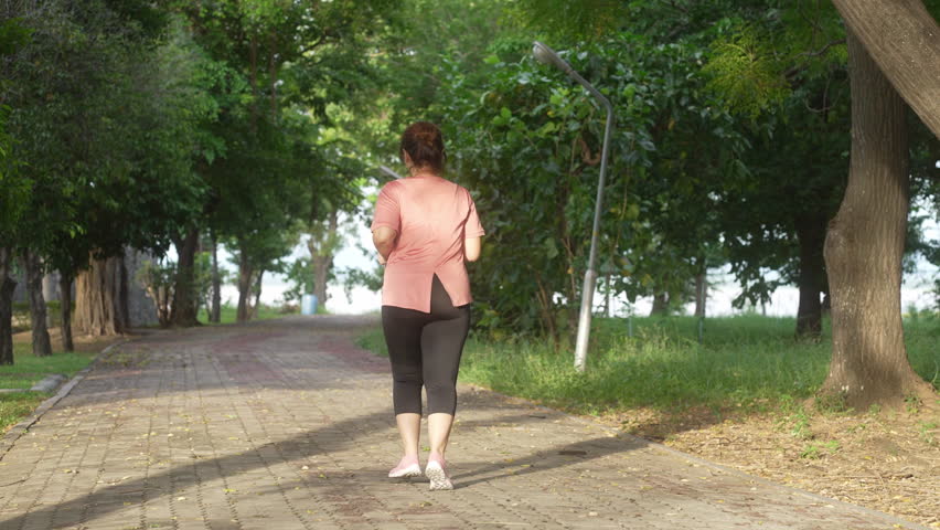 A middle-aged, overweight Asian woman jogging for exercise on a park road in the evening, dressed in athletic wear and holding a water bottle, symbolizing a commitment to a healthy lifestyle.