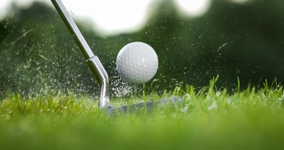 Golf club hits a golf ball in a super slow motion. Drops of morning dew and grass particles rise into the air after the impact.
