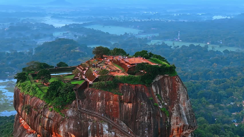 Multiple tourists walk by the ruins of the ancient stone fortress on the top of the picturesque mountain. Sigiriya palace, Sri Lanka, South Asia. Top view. Hazy scenery at backdrop.