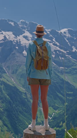 Vertical video. An attractive girl in a blue shirt and a fashionable hat spends her holidays alone, traveling through the Alpine meadows. Tourist with a backpack travels through Switzerland