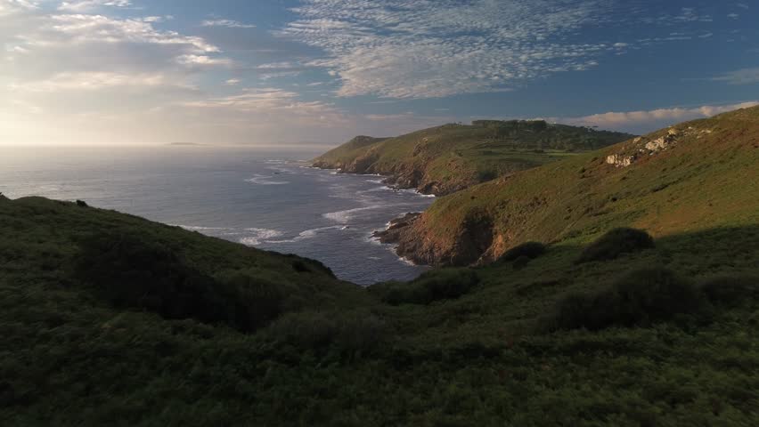 Gliding over calm Atlantic Ocean and rugged cliffs of the West of Ons Island. Verdant hills, with scattered trees, crossed by trails, meet rocky cliffs and crashing waves. Sálvora island in background