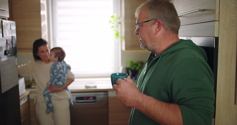 Man smiling and holding cup in foreground while mother holds baby in kitchen, cheerful family interaction during casual morning routine, home lifestyle