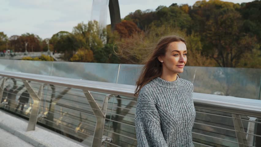 A young woman wearing a stylish sweater poses confidently on a modern bridge in an urban setting