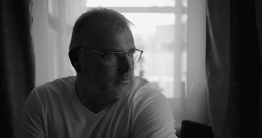 Black and white portrait of a man seated indoors, gazing thoughtfully to the side near a window, curtains and soft light creating a calm reflective atmosphere