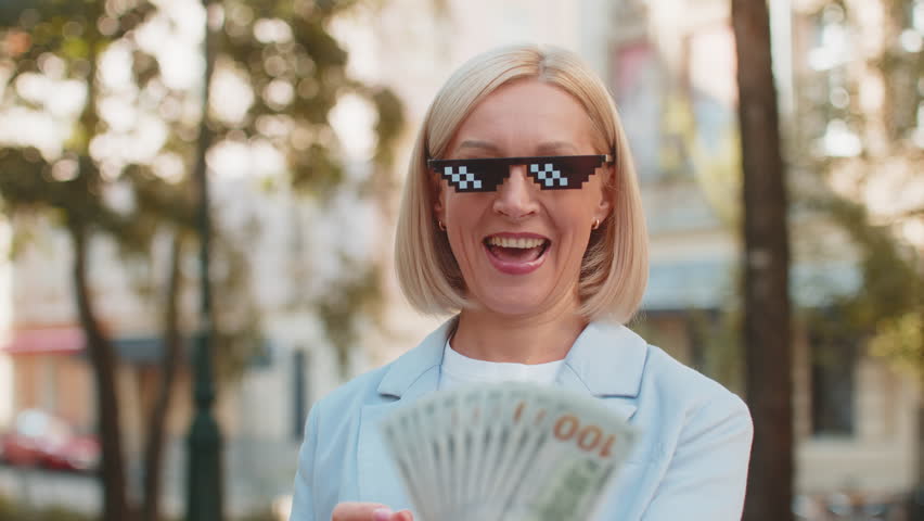 Happy rich Caucasian mature businesswoman putting on thug life sunglasses showing money dollar cash banknotes standing on city street. Success business career, lottery winner, big income, wealth.