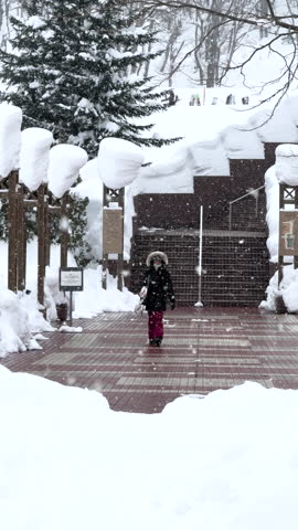 VERTICAL: Snowy entrance at ski resort with a snowboarder walking away after riding fresh powder snow. Large drifts of snow rest on wooden pillars and edges of stairs as smiling rider leaves the area.