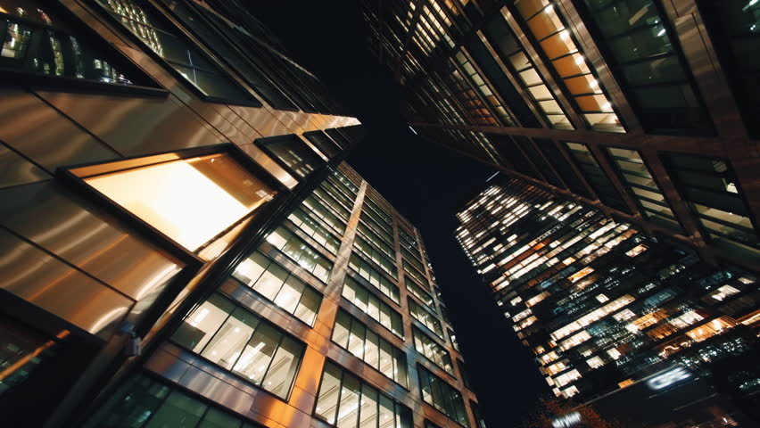 Wide angle gimbal dolly shot downtown high-rise glass skyscrapers at night, financial district looking up tall modern architecture office buildings city center street, warm lighted windows