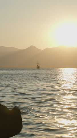 Vertical video. A Serene Sunset Over the Sea with Silhouetted Mountains in the Distance, a Small Boat Sailing Peacefully on the Golden-Reflecting Water. Slow motion.