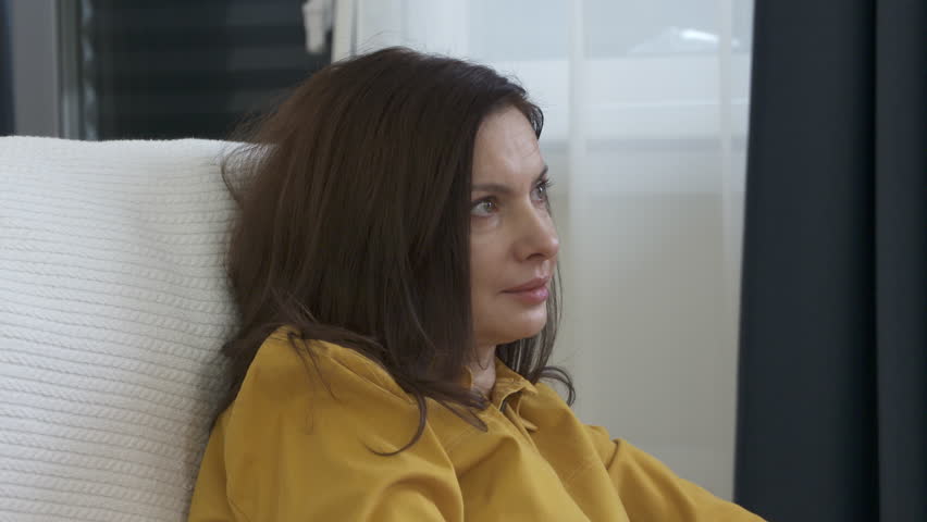 Adult 44 year old woman sitting on sofa in living room in front of TV watching melodrama. Woman with tear stained face smiles worried while watching movie.