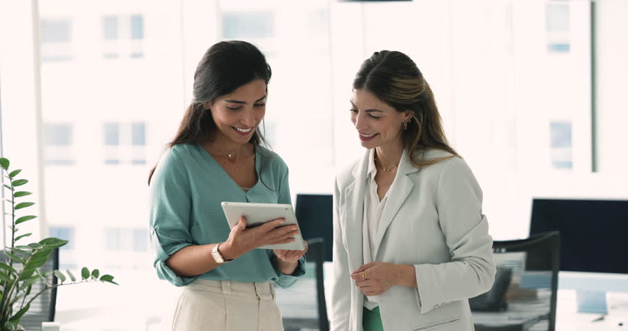 Two positive Hispanic female business coworkers discussing online content on tablet, holding gadget, looking at display. Manager woman showing electronic report, presentation to colleague for advice