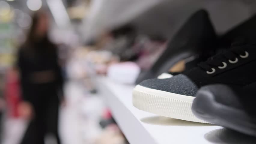 Row of Shoes on Store Shelf with Blurred Background of Shoppers in a Busy Footwear Retail Display.