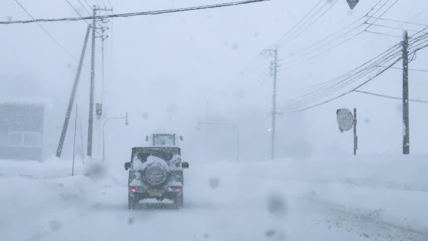 Snowy winter road with cars navigating through heavy snowfall and icy conditions. Traffic signs and power lines on the sides are blanketed with thick snow, roadway is surrounded by high snowdrifts.