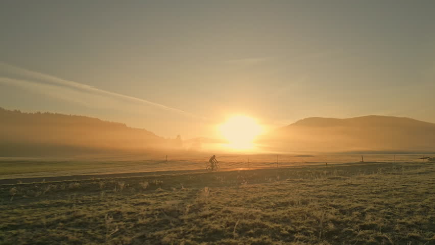 Man cycling through rural fields road during sunset, side view tracking shot. Cyclist silhouette riding bike in countryside scenery with mountains and sun on horizon, wide shot