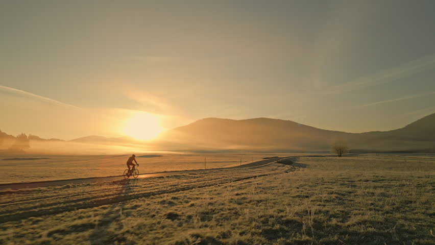 Man cycling through rural fields road during sunset, side view tracking shot. Cyclist silhouette riding bike in countryside scenery with mountains and sun on horizon, wide shot