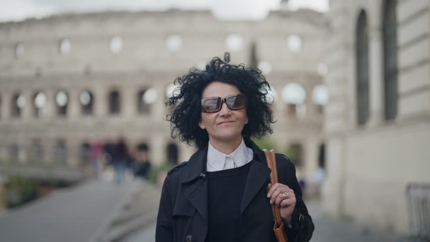 Smiling business woman walk with Colosseum in background in Rome, Italy, wearing a trench coat and sunglasses.