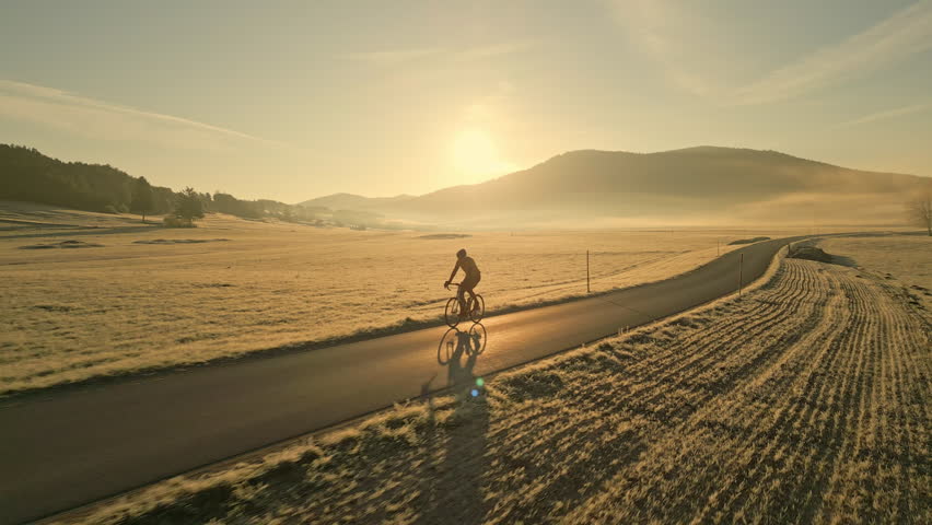 Cyclist riding along quiet rural road at sunset, wide tracking shot. Man cycling in peaceful countryside scene. Agricultural fields and golden hills landscape aerial view