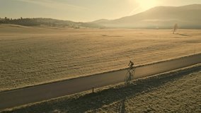 Male cyclist biking through picturesque countryside aerial tracking shot. Man cycling on empty road surrounded by misty golden fields at sunrise sun drone view - Powered by Shutterstock - Get 15% off with code: PIKWIZARD15