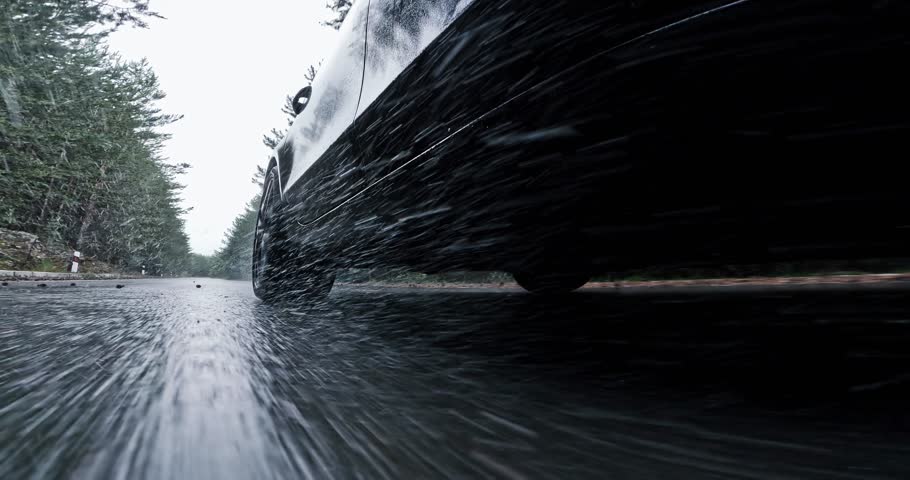 A car is driving down a wet road with rain coming down spraying water drops behind it toward camera