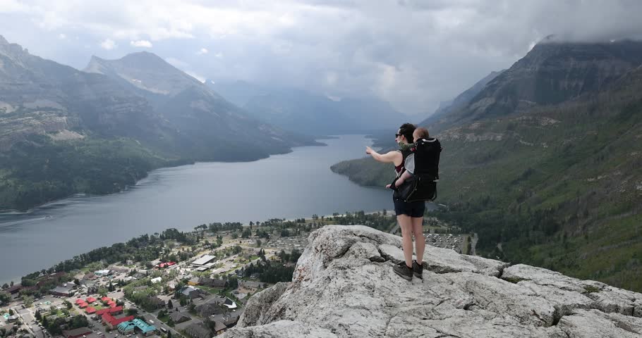 A mother hikes with her baby to the top of a mountain, pointing out to the amazing mountain and lake views. Waterton National Park, Alberta, Canada.