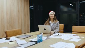A cheerful woman wearing a festive Santa hat is diligently working on her laptop in a modern office filled with scattered papers, women celebrating Christmas at work - Powered by Shutterstock - Get 15% off with code: PIKWIZARD15