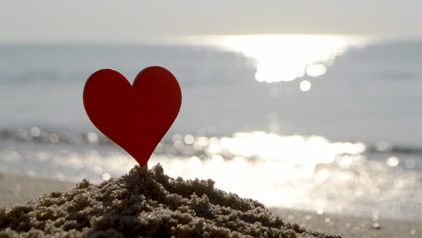 Plastic stick in shape of red heart in sand on sandy beach of sea shore on background of sea waves on sunny summer day close-up. Concept Love relationship romance amorousness amour St Valentines Day