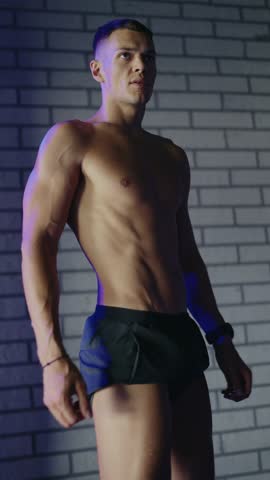 Strong muscular man posing shirtless in a gym, showcasing fit physique and determination. Bricks wall in background. Muscular Man Posing. Vertical video.