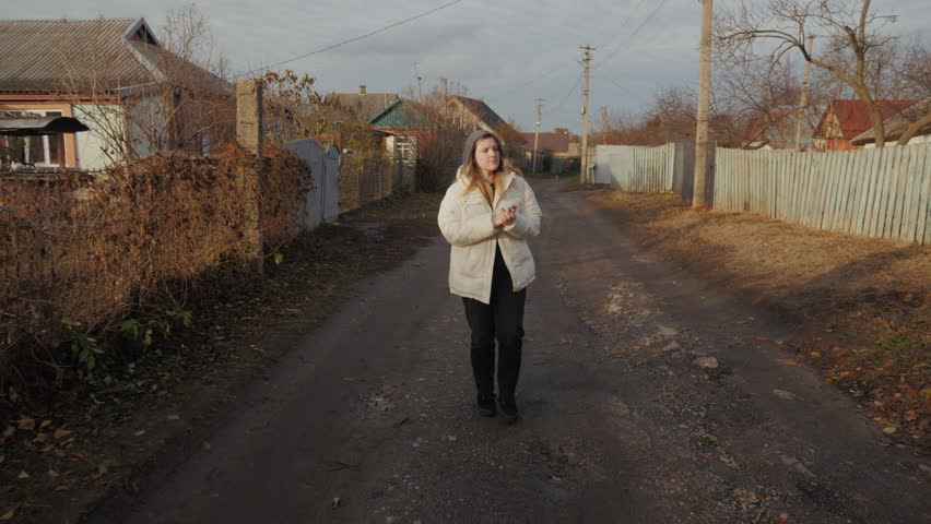 A young woman walks through a muddy alley bordered by rundown single-story houses, shivering in the cold and trying to warm her hands