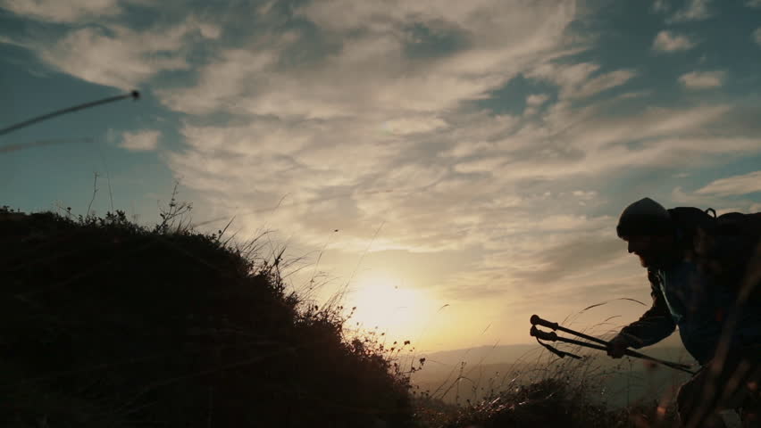 A hiker climbs a mountainside against a sunset backdrop. The footage is shot from the side, with only the dark silhouette of the figure visible against the bright sun. The atmosphere emphasizes a sens