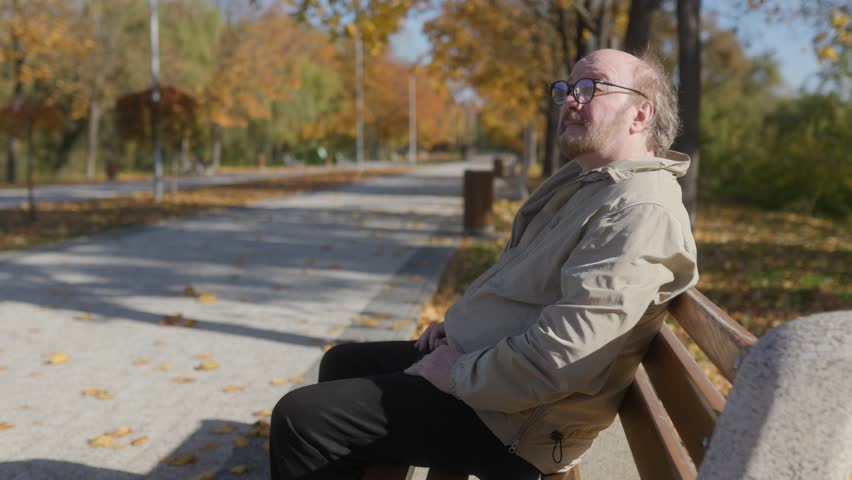 Old man with glasses and a beard finds solace on a park bench in autumn. The peaceful surroundings and vibrant foliage create a serene outdoor scene.